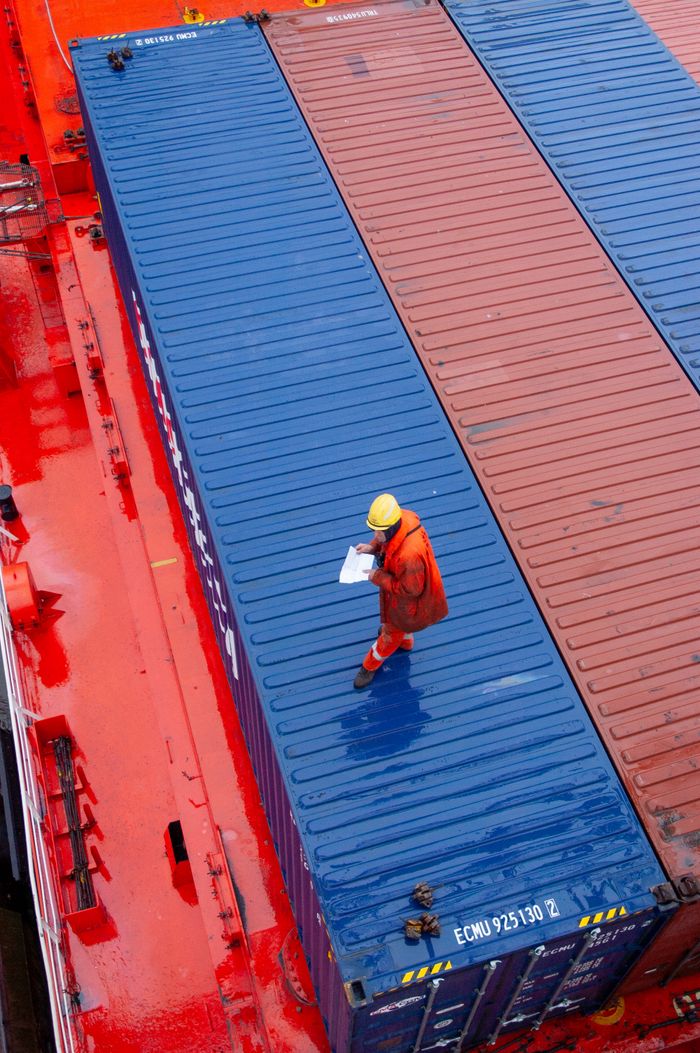 services-01 Overhead view of a worker inspecting containers on a ship deck in a maritime setting.