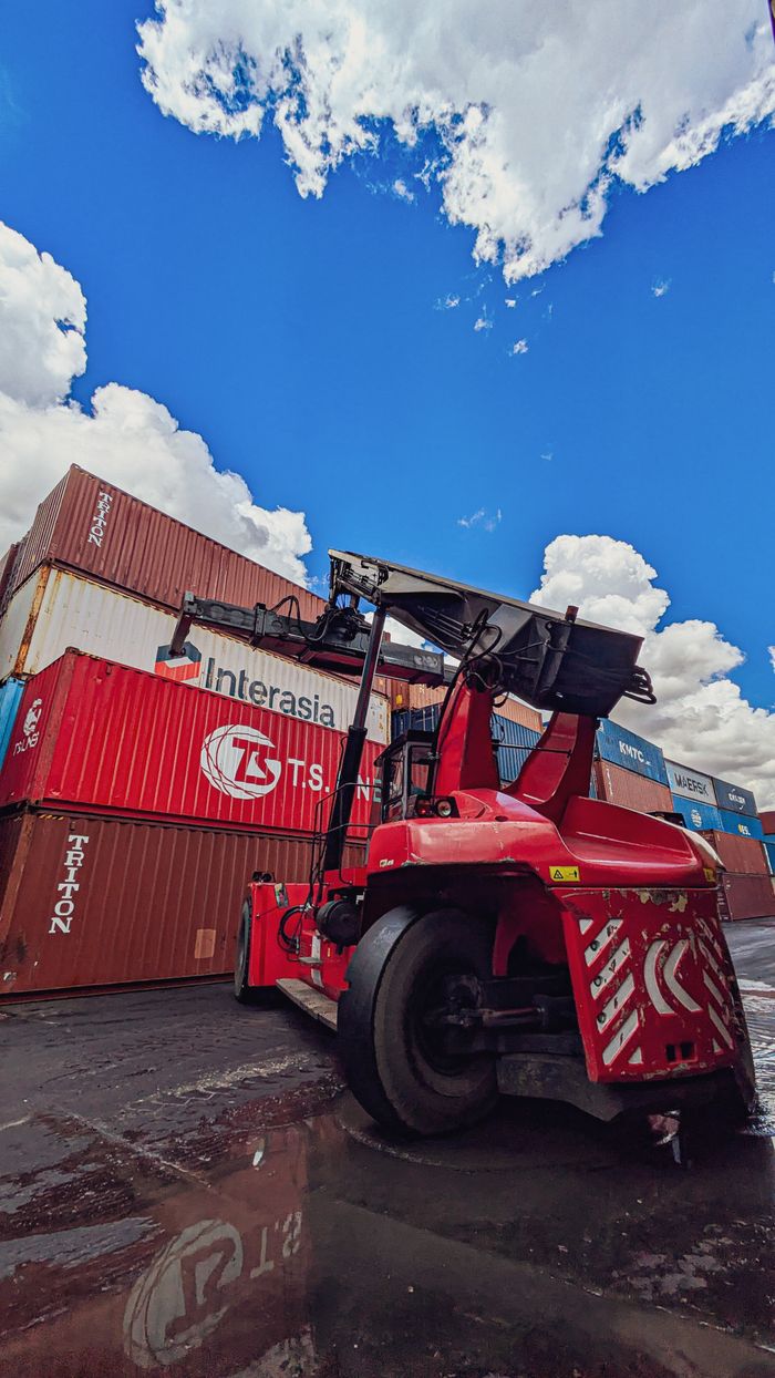 about-img Dynamic view of a red forklift amidst stacked shipping containers under a vibrant blue sky.